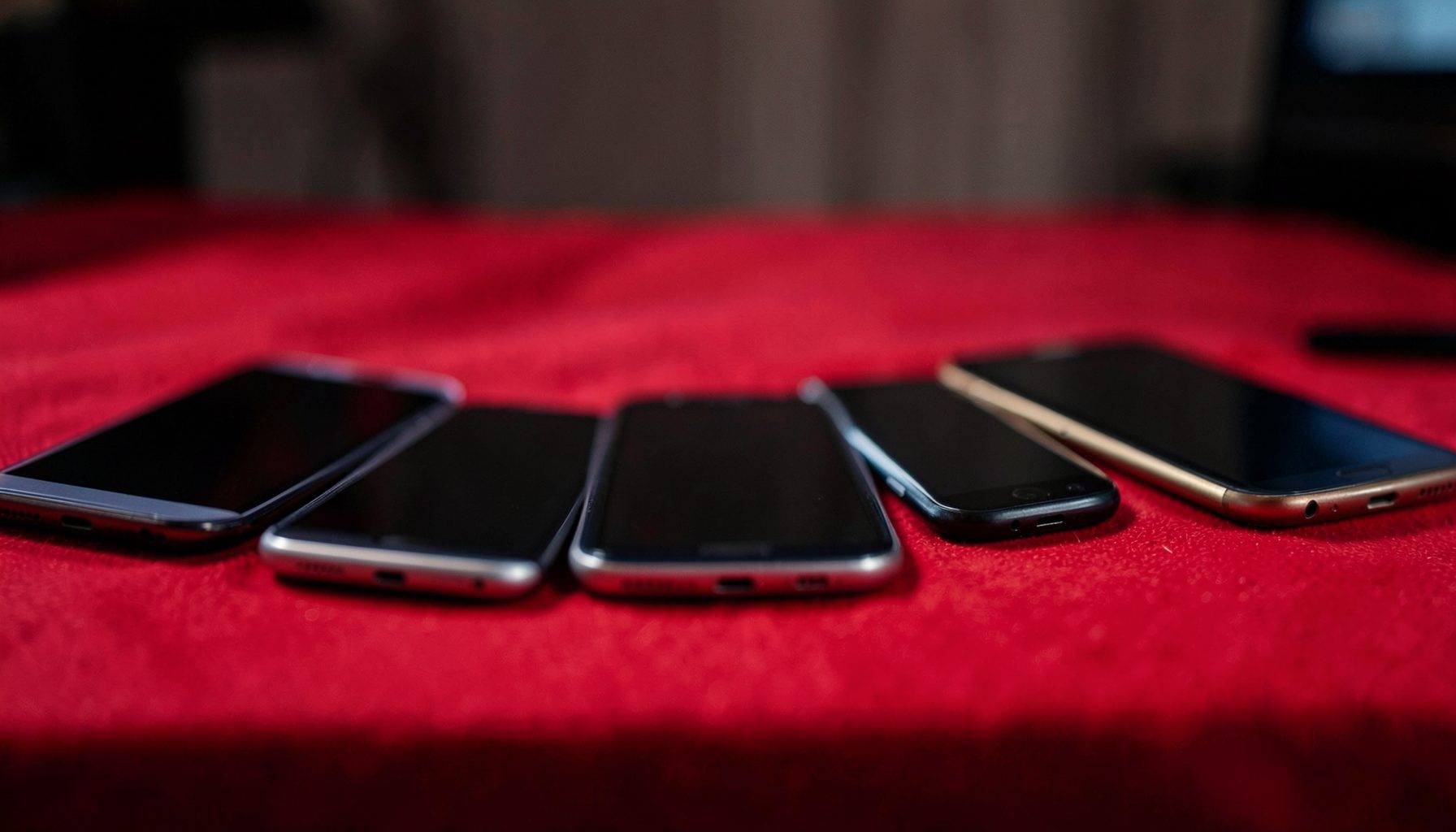 Deretan smartphone berbagai merek tersusun rapi di atas meja, layar mati, suasana ruangan remang-remang
