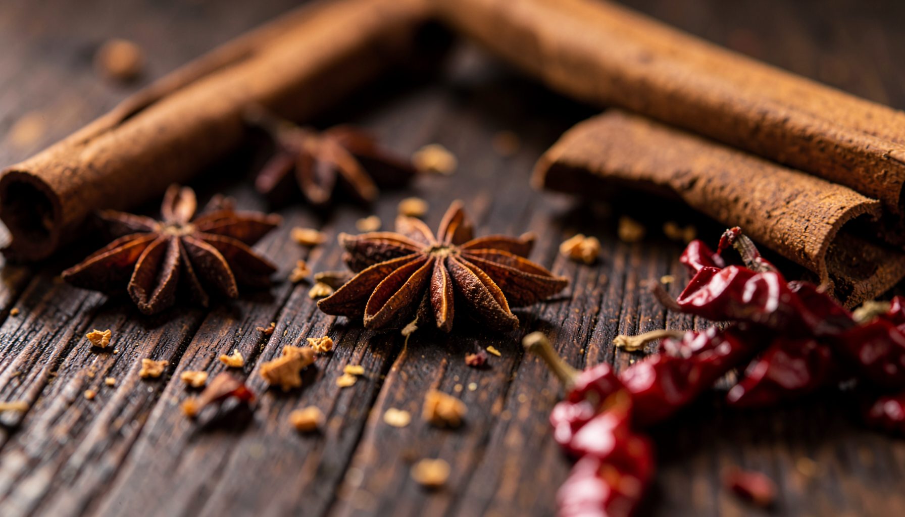 Close-up photograph of rich, dark wooden table surface with scattered aromatic spices, star anise, cinnamon sticks, and dried chili peppers, warm amber and deep brown tones, rustic culinary atmosphere